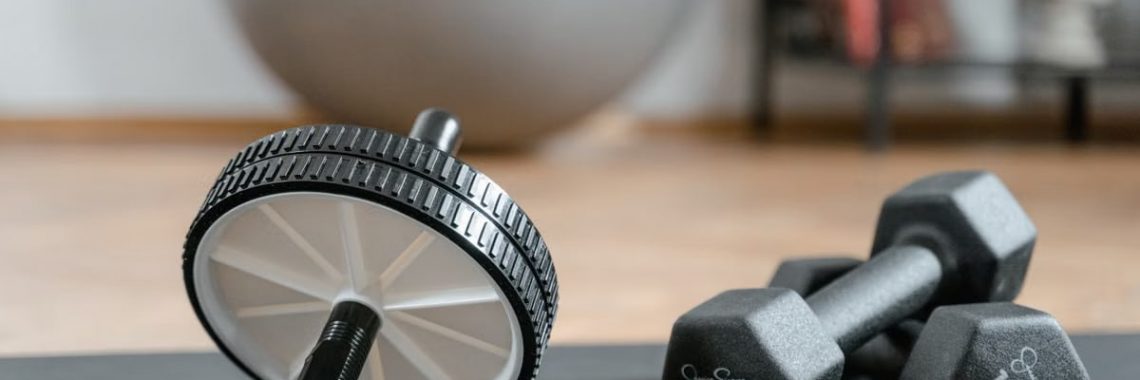 Close-up of ab wheel and dumbbells on a mat in a home gym setting.