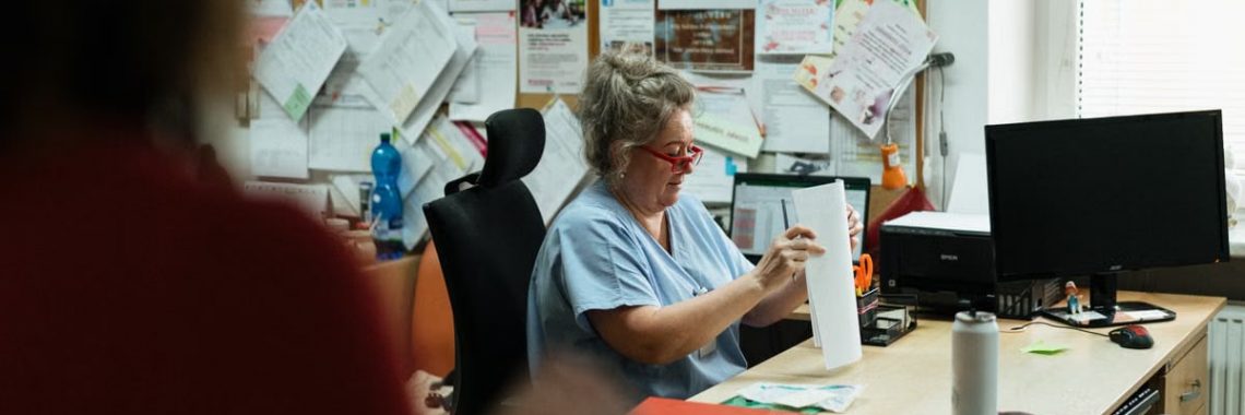 A senior caregiver in blue scrubs sorting paperwork at a hospital office desk.